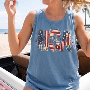 Könnte beinhalten: Ein blaues Tanktop in Jeansoptik mit dem Schriftzug USA in einer großen, dekorativen Schriftart. Die Buchstaben sind mit rot-weiß-blauen Mustern wie Sternen, Streifen, Vichy-Karos und Blumen gefüllt und mit kleinen Schleifen und Herzen verziert.