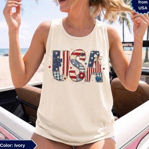 Könnte beinhalten: Ein elfenbeinfarbenes Tanktop mit dem Schriftzug USA in einer Patchwork-Grafik. Die Buchstaben sind mit roten, weißen und blauen Mustern verziert, darunter Sterne, Streifen und Vichy-Karos, akzentuiert durch kleine Schleifen, eine Blume und ein Herz.