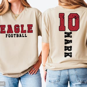May include: A sand-colored short-sleeve t-shirt featuring a distressed red and black graphic on the front that reads EAGLE FOOTBALL. The back of the shirt displays the number 10 in red and the name MARK in black, both in a distressed font style.
