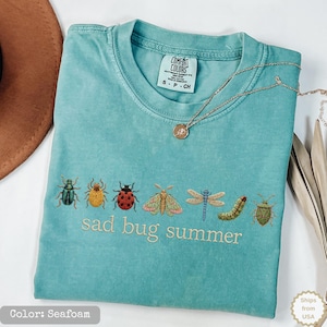May include: A seafoam green short-sleeve t-shirt featuring an embroidered row of various insects including a beetle, ladybug, moth, dragonfly, and caterpillar, with the text "sad bug summer" stitched in light thread below the insect graphic.