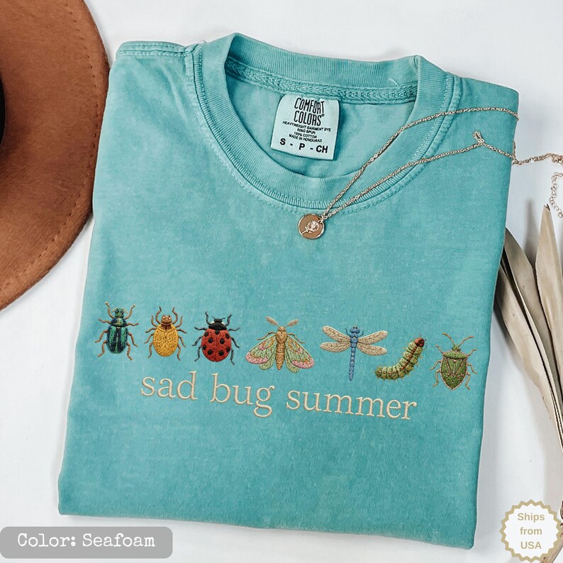 May include: A seafoam green short-sleeve t-shirt featuring an embroidered row of various insects including a beetle, ladybug, moth, dragonfly, and caterpillar, with the text "sad bug summer" stitched in light thread below the insect graphic.