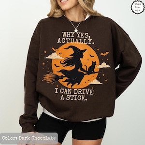 Può includere: Una felpa a maniche lunghe color marrone cioccolato scuro con la grafica di una strega su una scopa e un gatto davanti a una grande luna arancione. Il testo recita: WHY YES, ACTUALLY. I CAN DRIVE A STICK. Il design include piccole stelle e nuvole.