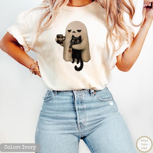 May include: An ivory t-shirt featuring a graphic of a beige ghost holding a black cat and a dark mug with the text MEH printed on it. The ghost has a sad facial expression, and the black cat is holding a small orange pumpkin charm.