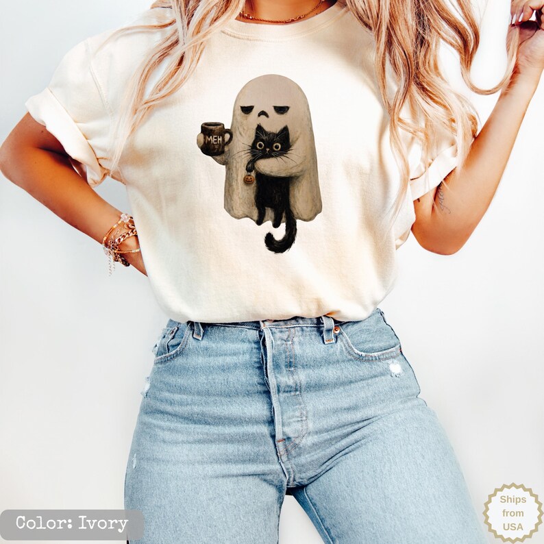 May include: An ivory t-shirt featuring a graphic of a beige ghost holding a black cat and a dark mug with the text MEH printed on it. The ghost has a sad facial expression, and the black cat is holding a small orange pumpkin charm.