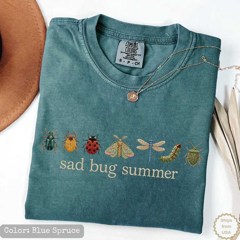 May include: A blue spruce t-shirt featuring a row of embroidered insects including a beetle, ladybug, moth, dragonfly, and caterpillar. The text "sad bug summer" is embroidered in light beige below the insects. The shirt has a Comfort Colors label.