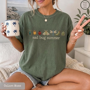 May include: A moss green crewneck t-shirt featuring a graphic of various insects including a ladybug, moth, and dragonfly, with the text "sad bug summer" printed in a simple font. The shirt is paired with a white mug decorated with blue palm trees.