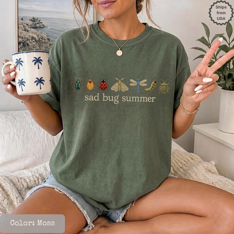May include: A moss green crewneck t-shirt featuring a graphic of various insects including a ladybug, moth, and dragonfly, with the text "sad bug summer" printed in a simple font. The shirt is paired with a white mug decorated with blue palm trees.