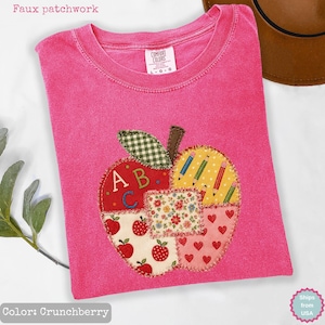 Könnte beinhalten: Ein leuchtend pinkfarbenes Kurzarm-T-Shirt mit einem großen Apfel-Design in Faux-Patchwork-Optik auf der Vorderseite. Die Apfelgrafik enthält Abschnitte mit roten Äpfeln, Herzen, Stiften sowie den Buchstaben A, B und C, die mit Ziernähten versehen sind.