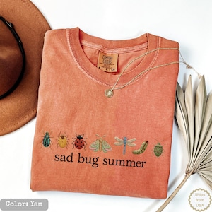 May include: A yam-colored crew neck t-shirt featuring a row of embroidered insects including beetles, a moth, a dragonfly, and a caterpillar above the black text "sad bug summer". The shirt has a Comfort Colors brand label at the inner neckline.