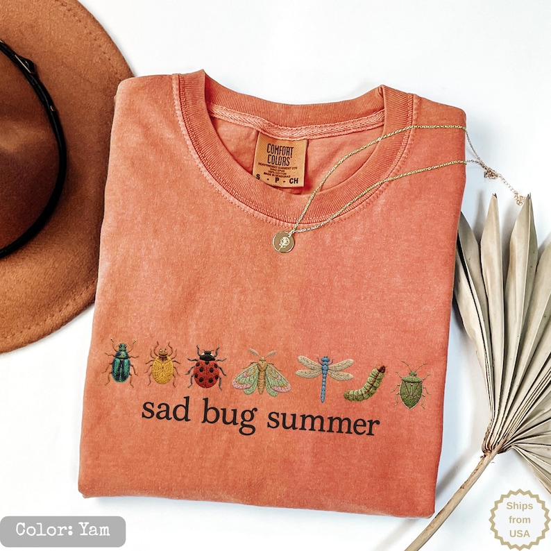 May include: A yam-colored crew neck t-shirt featuring a row of embroidered insects including beetles, a moth, a dragonfly, and a caterpillar above the black text "sad bug summer". The shirt has a Comfort Colors brand label at the inner neckline.