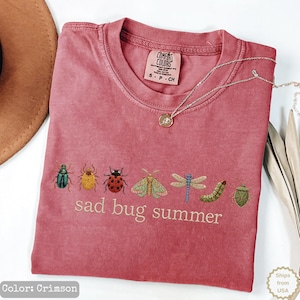 May include: A crimson short-sleeved t-shirt featuring an embroidered row of various colorful insects including a beetle, ladybug, moth, dragonfly, and caterpillar, with the text "sad bug summer" printed in a light beige font below the insects.