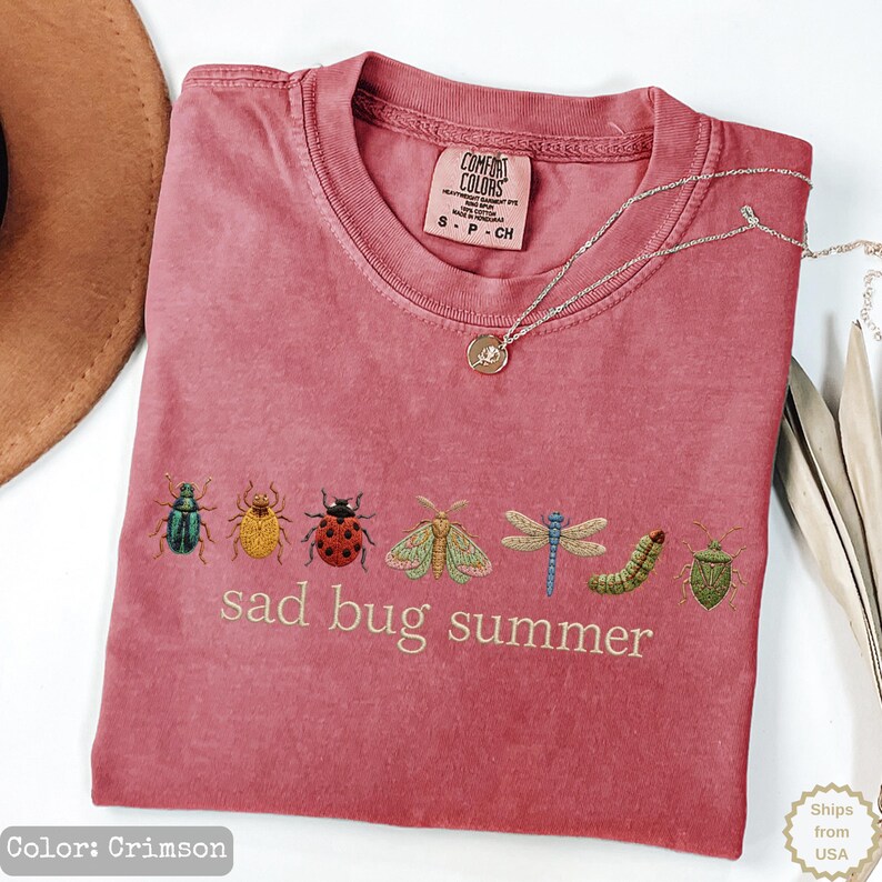 May include: A crimson short-sleeved t-shirt featuring an embroidered row of various colorful insects including a beetle, ladybug, moth, dragonfly, and caterpillar, with the text "sad bug summer" printed in a light beige font below the insects.