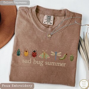 May include: An espresso-colored t-shirt featuring a row of various insect illustrations and the text "sad bug summer" printed in white. The shirt has a crew neckline and a Comfort Colors brand label inside the collar. A silver necklace rests on top.