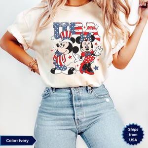 May include: Ivory-colored t-shirt featuring a patriotic design with the word "USA" in red, white, and blue. The graphic includes Mickey and Minnie Mouse dressed in patriotic attire, with stars and stripes. A fun and festive design for the 4th of July.