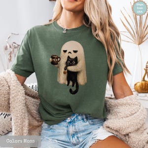 May include: A moss green t-shirt featuring a graphic of a white ghost holding a brown mug with the text MEH and a black cat. The ghost has a neutral expression, and the cat has large eyes and a small pumpkin charm on its collar.