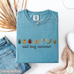 May include: A blue short-sleeved t-shirt featuring an embroidered row of various colorful insects including a ladybug, dragonfly, and moth, with the text "sad bug summer" printed in black lettering below the insects and a silver chain necklace.
