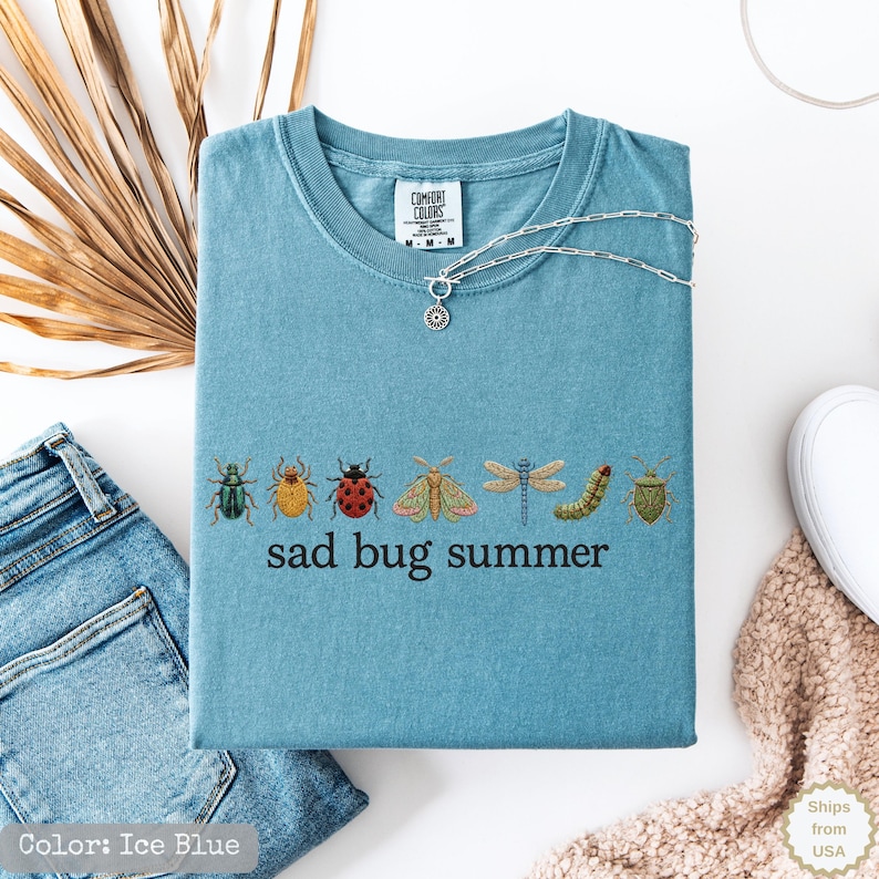 May include: A blue short-sleeved t-shirt featuring an embroidered row of various colorful insects including a ladybug, dragonfly, and moth, with the text "sad bug summer" printed in black lettering below the insects and a silver chain necklace.