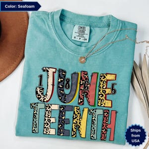 Op de afbeelding: Zeegroen t-shirt met de tekst "JUNE TEENTH" in een kleurrijk luipaardprint ontwerp. Het shirt heeft een ronde hals en korte mouwen. Een gouden ketting met een hanger is over het shirt gedrapeerd.