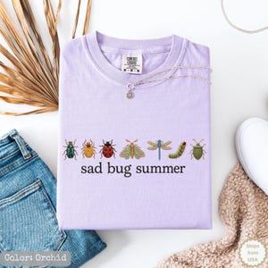May include: A light purple t-shirt featuring a row of seven colorful insect illustrations including beetles, a moth, a dragonfly, and a caterpillar, with the black text "sad bug summer" printed underneath the insects on the front of the garment.