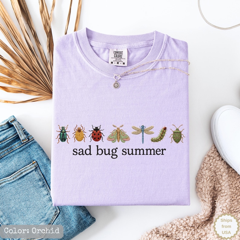 May include: A light purple t-shirt featuring a row of seven colorful insect illustrations including beetles, a moth, a dragonfly, and a caterpillar, with the black text "sad bug summer" printed underneath the insects on the front of the garment.