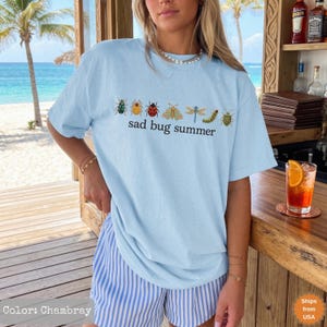 May include: A light blue t-shirt featuring a row of colorful insect illustrations including a beetle, ladybug, moth, dragonfly, and caterpillar, with the black text "sad bug summer" printed underneath the insects on the front of the garment.