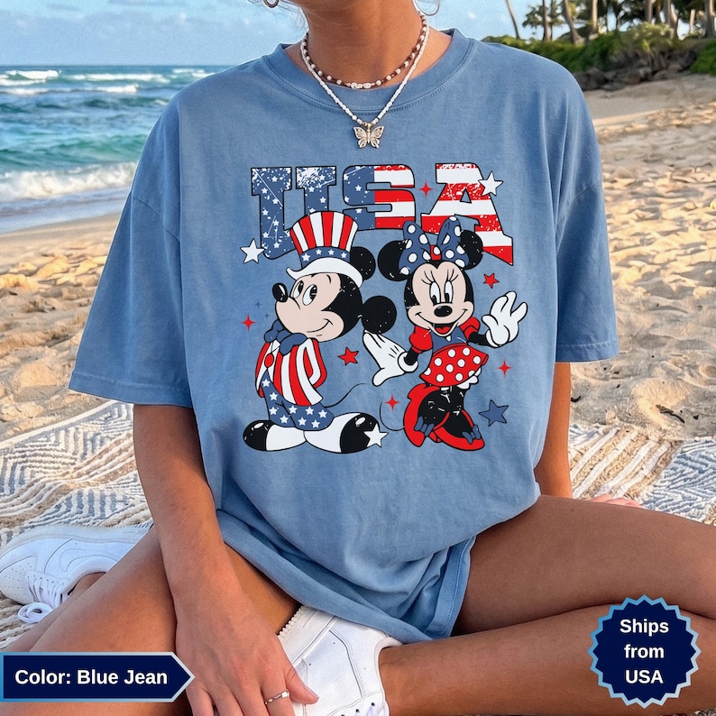 May include: Blue jean t-shirt featuring Mickey and Minnie Mouse in patriotic attire with the text "USA". Mickey wears a hat and suit with red, white, and blue stripes and stars. Minnie wears a red and white polka dot skirt and bow.
