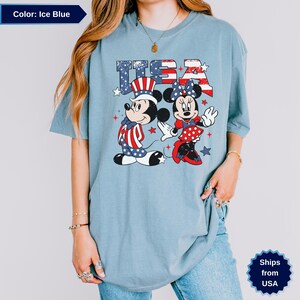 May include: Ice blue t-shirt featuring Mickey and Minnie Mouse in patriotic outfits with the text "USA". Mickey wears a red, white, and blue outfit and hat. Minnie wears a red polka dot dress and bow.