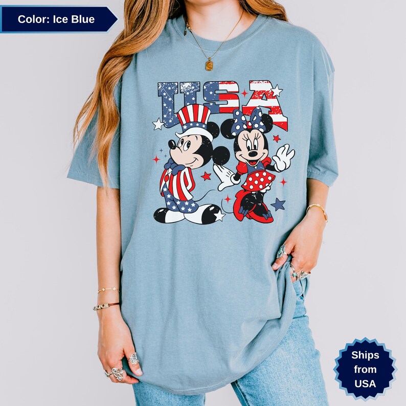 May include: Ice blue t-shirt featuring Mickey and Minnie Mouse in patriotic outfits with the text "USA". Mickey wears a red, white, and blue outfit and hat. Minnie wears a red polka dot dress and bow.