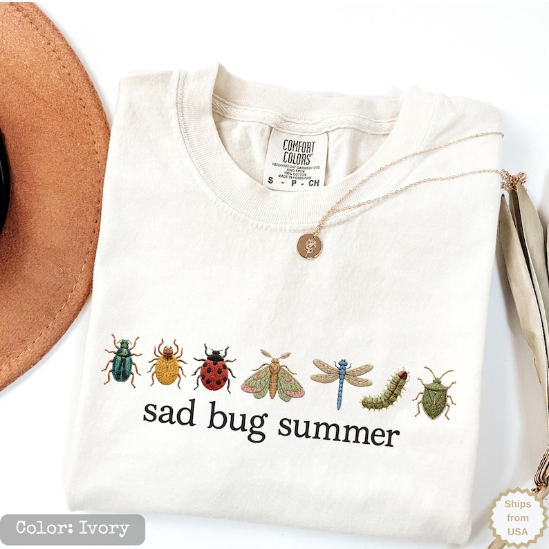 May include: An ivory t-shirt featuring a graphic of seven colorful insects arranged in a horizontal row above the black text "sad bug summer". The shirt has a crew neckline and a Comfort Colors brand label printed on the inside of the collar.