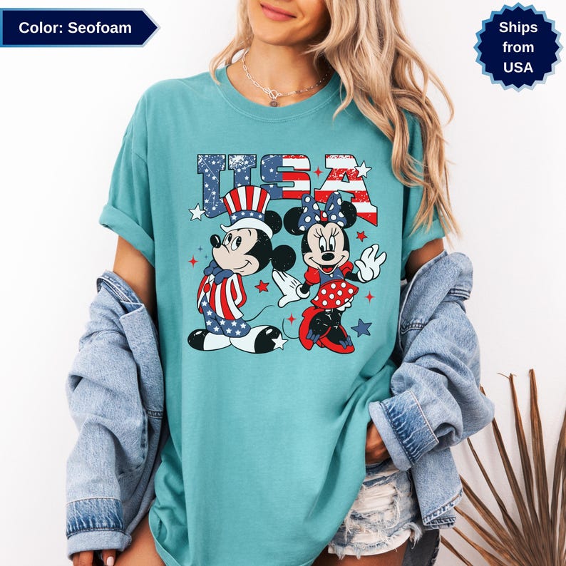 May include: Seofoam-colored t-shirt featuring Mickey and Minnie Mouse in patriotic attire. The word "USA" is printed above the characters in red, white, and blue. Mickey wears a top hat and Minnie a bow, both in patriotic colors.