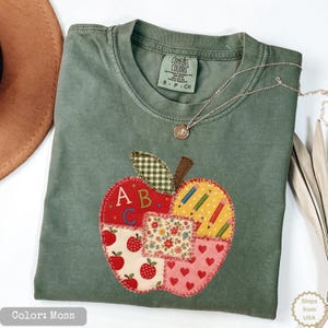 Könnte beinhalten: Ein moosgrünes Kurzarm-T-Shirt mit einer großen, gemusterten Apfel-Applikation auf der Vorderseite. Das Apfeldesign besteht aus Abschnitten mit roten Äpfeln, Blumenmustern, roten Herzen und Stiften sowie den Buchstaben A, B und C im oberen Bereich.