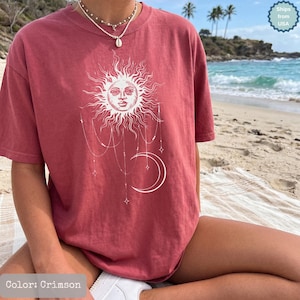Puede incluir: Una camiseta de manga corta color carmesí con un estampado gráfico blanco de un sol con rostro humano, una luna creciente y cadenas de cuentas colgantes con detalles de estrellas. La prenda tiene cuello redondo y corte holgado.