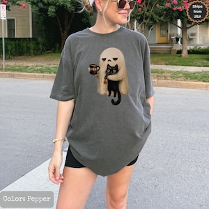 May include: A charcoal gray short-sleeve t-shirt featuring a graphic print of a beige ghost holding a brown mug with the text MEH and a black cat with large eyes. The shirt has a crew neckline and a relaxed fit, showcasing a casual style.