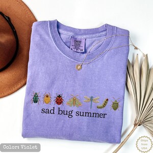 May include: A violet short-sleeve t-shirt featuring an embroidered row of various insects including a beetle, ladybug, moth, dragonfly, and caterpillar, with the text "sad bug summer" printed in black below the insects on the front of the shirt.