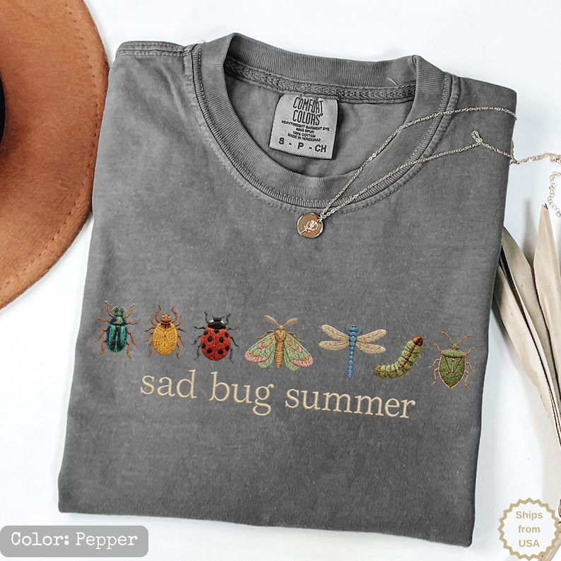 May include: A pepper-colored t-shirt featuring an embroidered row of various insects including a beetle, ladybug, moth, dragonfly, and caterpillar, with the text "sad bug summer" printed in a light beige font below the insect illustrations.
