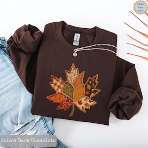 Könnte beinhalten: Ein dunkel schokoladenbraunes Sweatshirt mit Rundhalsausschnitt und einer großen, zentralen Ahornblatt-Applikation. Das Blatt besteht aus verschiedenen Stoffflicken in Orange-, Rot-, Grün- und Gelbtönen mit unterschiedlichen Mustern.