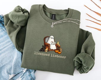 May include: A military green crewneck sweatshirt featuring a graphic of a ghost wearing glasses, sitting in a brown armchair and reading a book. Beside the chair are a black cat, a skull, candles, and pumpkins. The text reads licensed listener.
