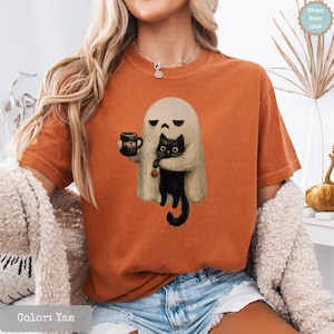 May include: A burnt orange short-sleeve t-shirt featuring a graphic print of a beige ghost holding a dark brown mug with the text "MEH" printed on it. The ghost is embracing a black cat that is holding a small orange pumpkin by its tail.