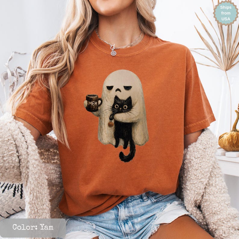 May include: A burnt orange short-sleeve t-shirt featuring a graphic print of a beige ghost holding a dark brown mug with the text "MEH" printed on it. The ghost is embracing a black cat that is holding a small orange pumpkin by its tail.