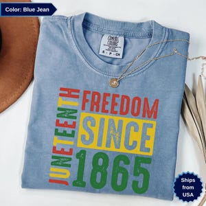 Op de afbeelding: Blauw spijkerbroekkleurig T-shirt met de woorden "JUNETEENTH FREEDOM SINCE 1865" in rood, geel en groen. Het shirt is van het merk Comfort Colors. Een gouden ketting ligt over het shirt.