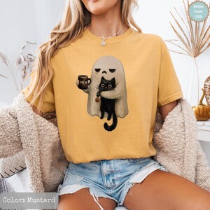 May include: A mustard yellow short-sleeve t-shirt featuring a graphic of a ghost with a grumpy expression holding a brown mug with the text MEH printed on it. The ghost is embracing a black cat that has a small orange pumpkin charm on its collar.