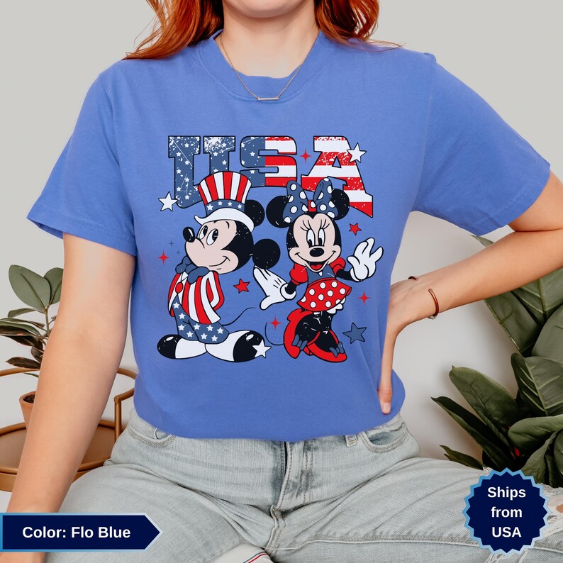 May include: Flo blue t-shirt featuring Mickey and Minnie Mouse in patriotic attire with the text "USA". Mickey Mouse wears a hat and suit with red, white, and blue stars and stripes. Minnie Mouse wears a polka dot skirt and bow.