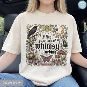 以下が含まれることがあります： アイボリー色のTシャツで、I find your lack of whimsy disturbingというテキストがプリントされています。デザインには、黒いカラス、蛾、三日月、甲虫、カタツムリ、キノコ、花のつるが落ち着いたアースカラーで描かれています。