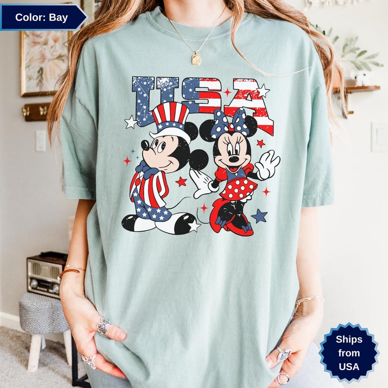 May include: A light blue t-shirt featuring Mickey and Minnie Mouse in patriotic outfits with the word "USA" in red, white, and blue. Mickey wears a top hat and Minnie has a bow. Stars and stripes complete the design.