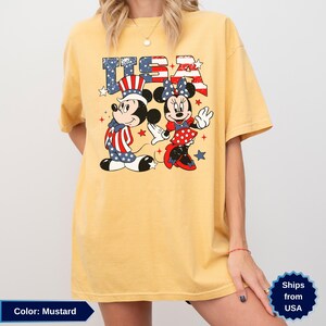 May include: Mustard-colored t-shirt featuring Mickey and Minnie Mouse in patriotic outfits with the word "USA" in a star-spangled design. Mickey wears a hat and vest with red, white, and blue stripes and stars. Minnie wears a polka-dot dress.