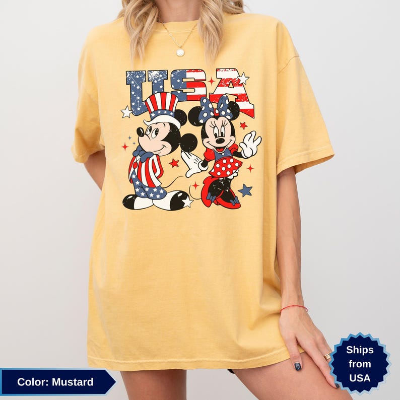 May include: Mustard-colored t-shirt featuring Mickey and Minnie Mouse in patriotic outfits with the word "USA" in a star-spangled design. Mickey wears a hat and vest with red, white, and blue stripes and stars. Minnie wears a polka-dot dress.