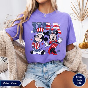 May include: Violet-colored t-shirt featuring Mickey and Minnie Mouse in patriotic attire with the text "USA". The design incorporates red, white, and blue elements, including stars and stripes, creating a festive look.