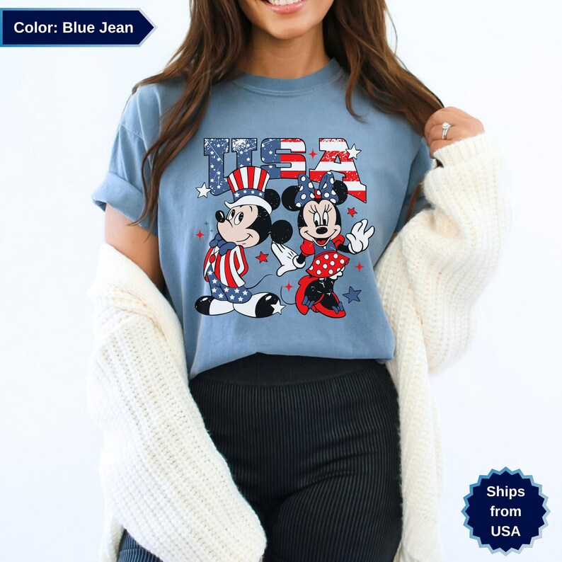 May include: Blue jean t-shirt featuring Mickey and Minnie Mouse in patriotic attire. The word "USA" is printed above the characters in red, white, and blue. Mickey Mouse is wearing a Uncle Sam hat and Minnie Mouse is wearing a polka dot skirt.