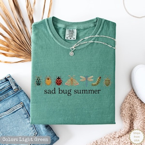 May include: A light green t-shirt featuring a row of various colorful insect illustrations including beetles, a moth, a dragonfly, and a caterpillar, with the text "sad bug summer" printed in black below the insects and a silver chain necklace.