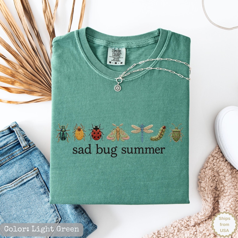 May include: A light green t-shirt featuring a row of various colorful insect illustrations including beetles, a moth, a dragonfly, and a caterpillar, with the text "sad bug summer" printed in black below the insects and a silver chain necklace.